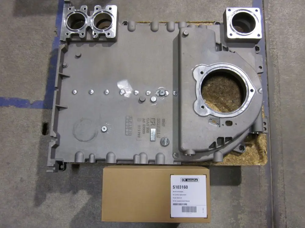 Produktbild eines Remeha Seitenglieds, metallisch, mit verschiedenen Öffnungen und Schrauben. Dazu ein brauner Karton mit Seriennummer, auf einem grauen Untergrund platziert.